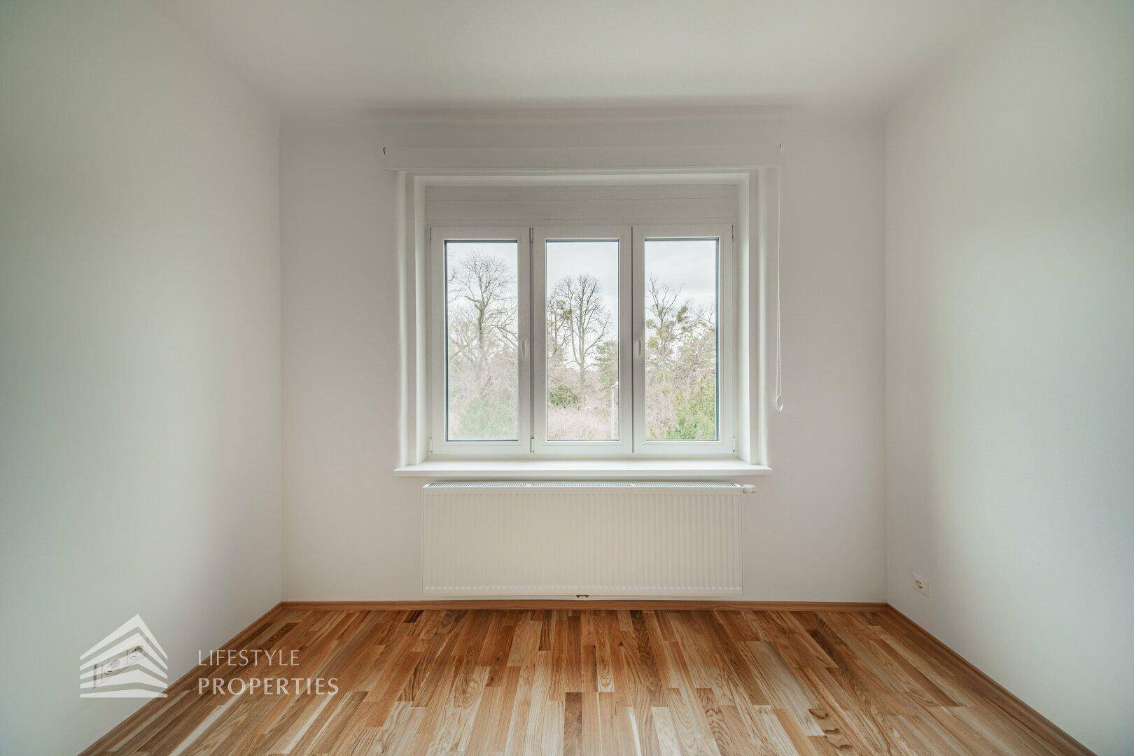 Helles Zimmer mit Parkettboden und einem großen Fenster, das eine grüne Aussicht bietet.