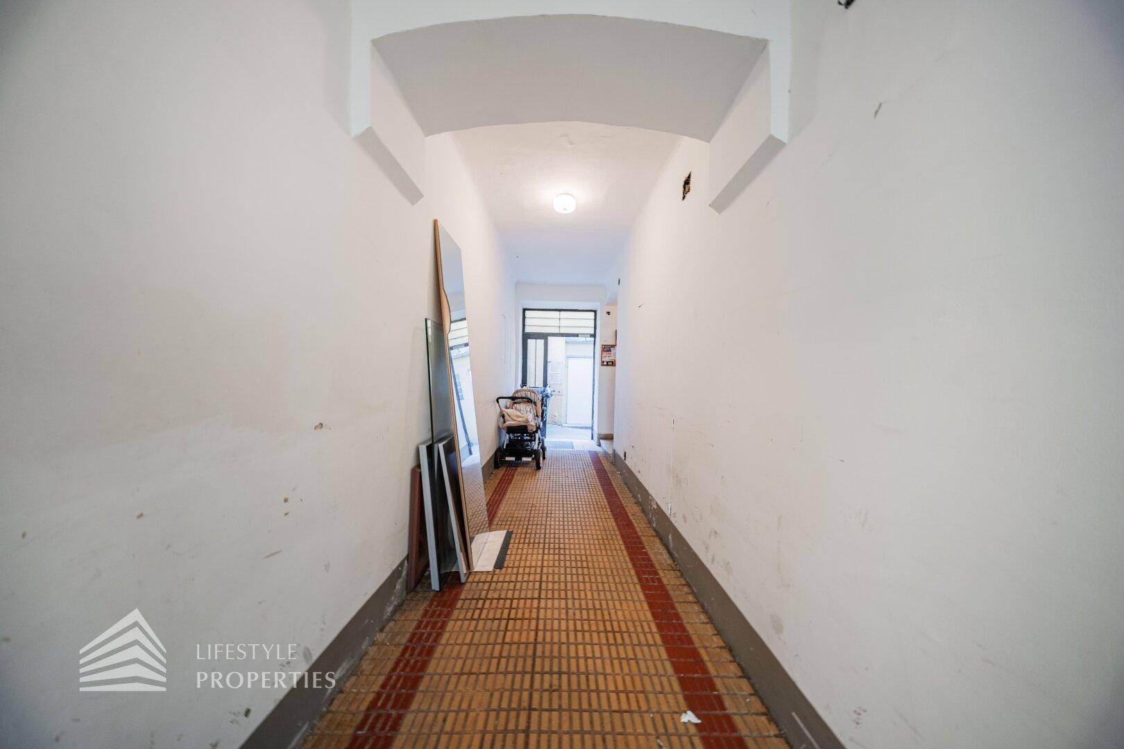 Schmaler, langer Flur mit hellen Wänden und einem gemusterten Fliesenboden, der zum Ausgang führt.