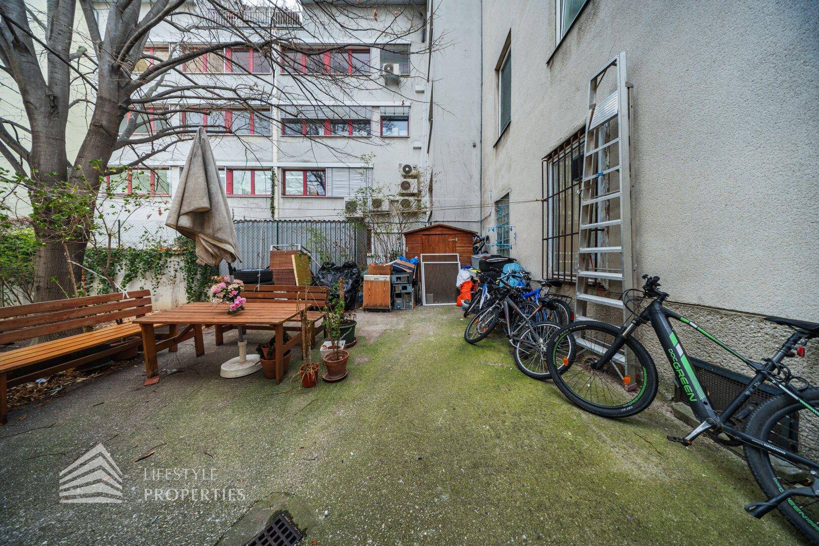 Hinterhofbereich mit einem Holztisch, Bänken und einem Sonnenschirm, umgeben von Gebäuden.