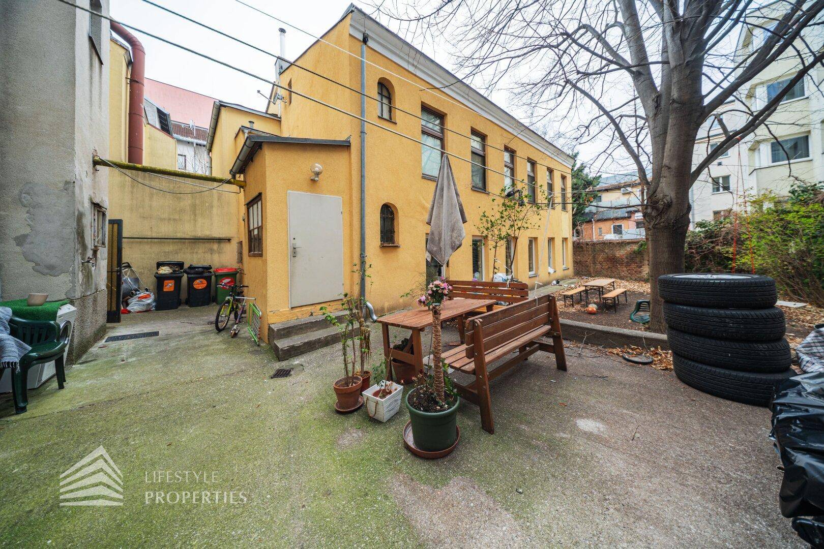 Geräumiger Hinterhof mit Sitzgelegenheiten, Bäumen und einer gelben Gebäudefassade im Hintergrund.