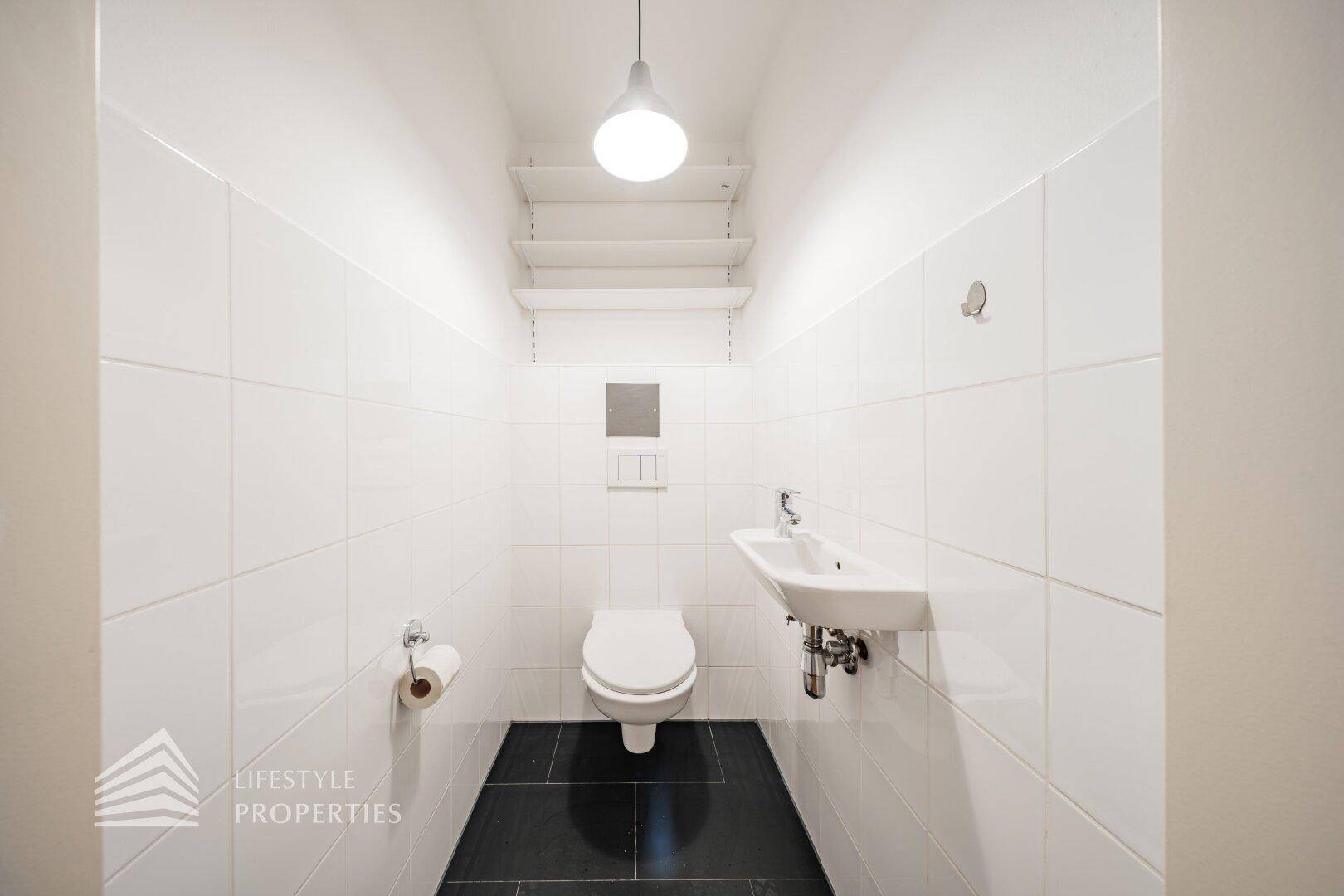 Separate Toilette mit wandhängendem WC, kleinem Waschbecken und weißen Fliesen.