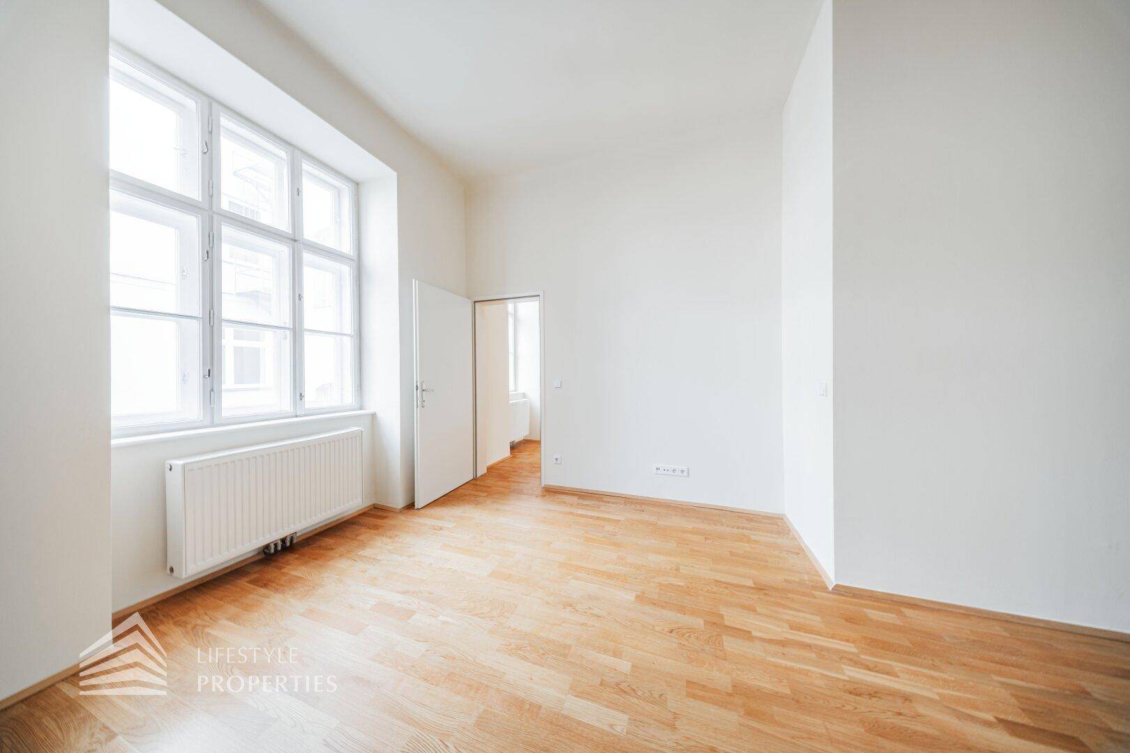 Geräumiges Zimmer mit großen Fenstern, hellem Parkettboden und viel Tageslicht.