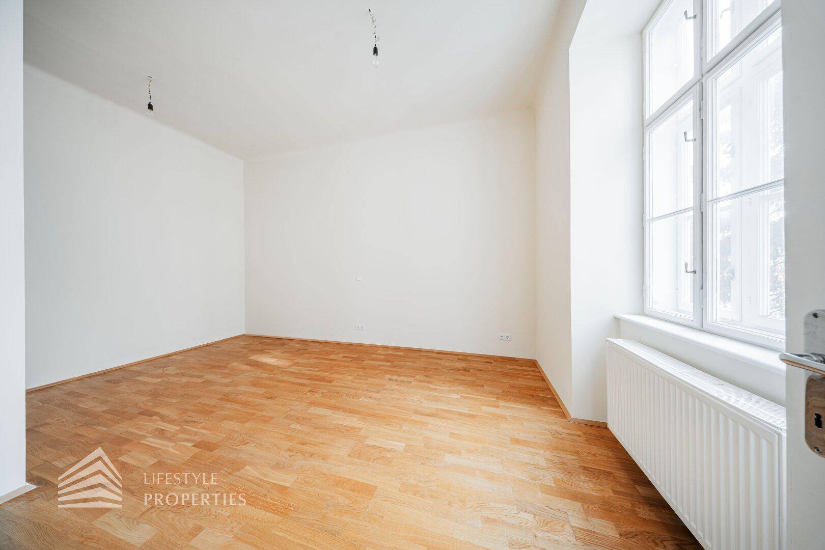 Leeres Zimmer mit hellem Parkettboden, weißen Wänden und großem Fenster.
