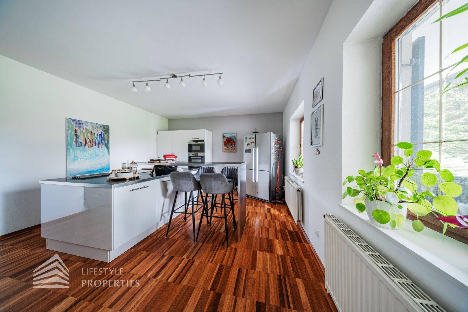 Helle Küche mit Kücheninsel, Holzboden und großem Fenster mit Blick ins Grüne.