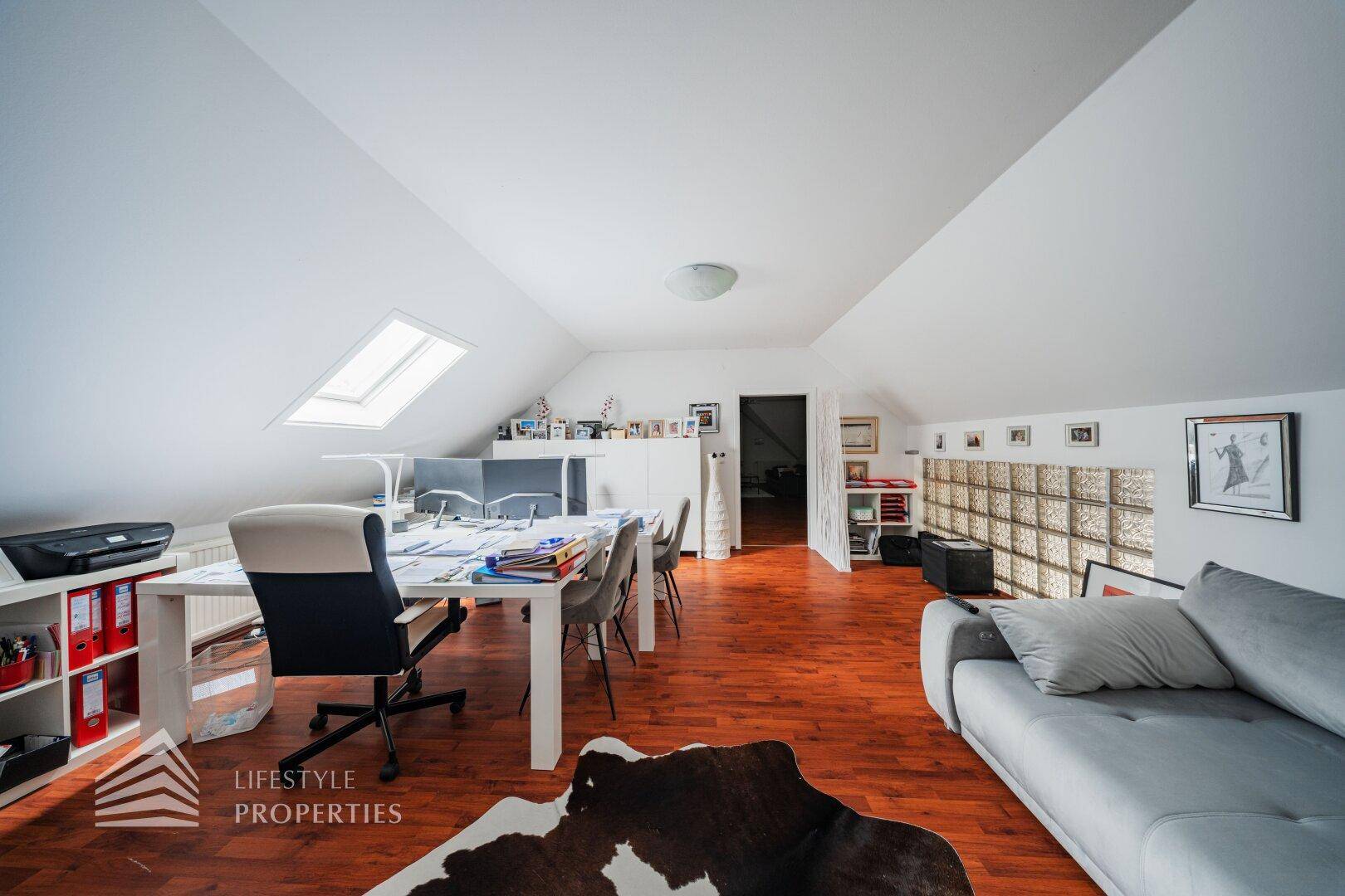 Geräumiges Dachgeschoss-Büro mit Holzboden, Sofa und praktischen Stauraummöglichkeiten.