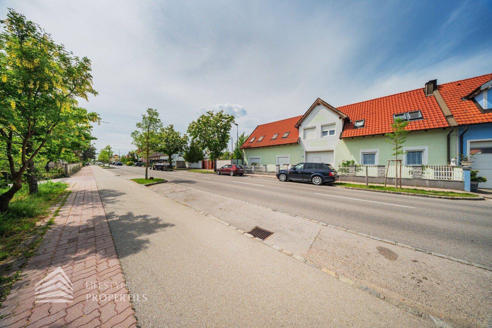Straßenansicht mit Bürgersteig, Fahrbahn und Wohnhäusern unter einem hellen Himmel.