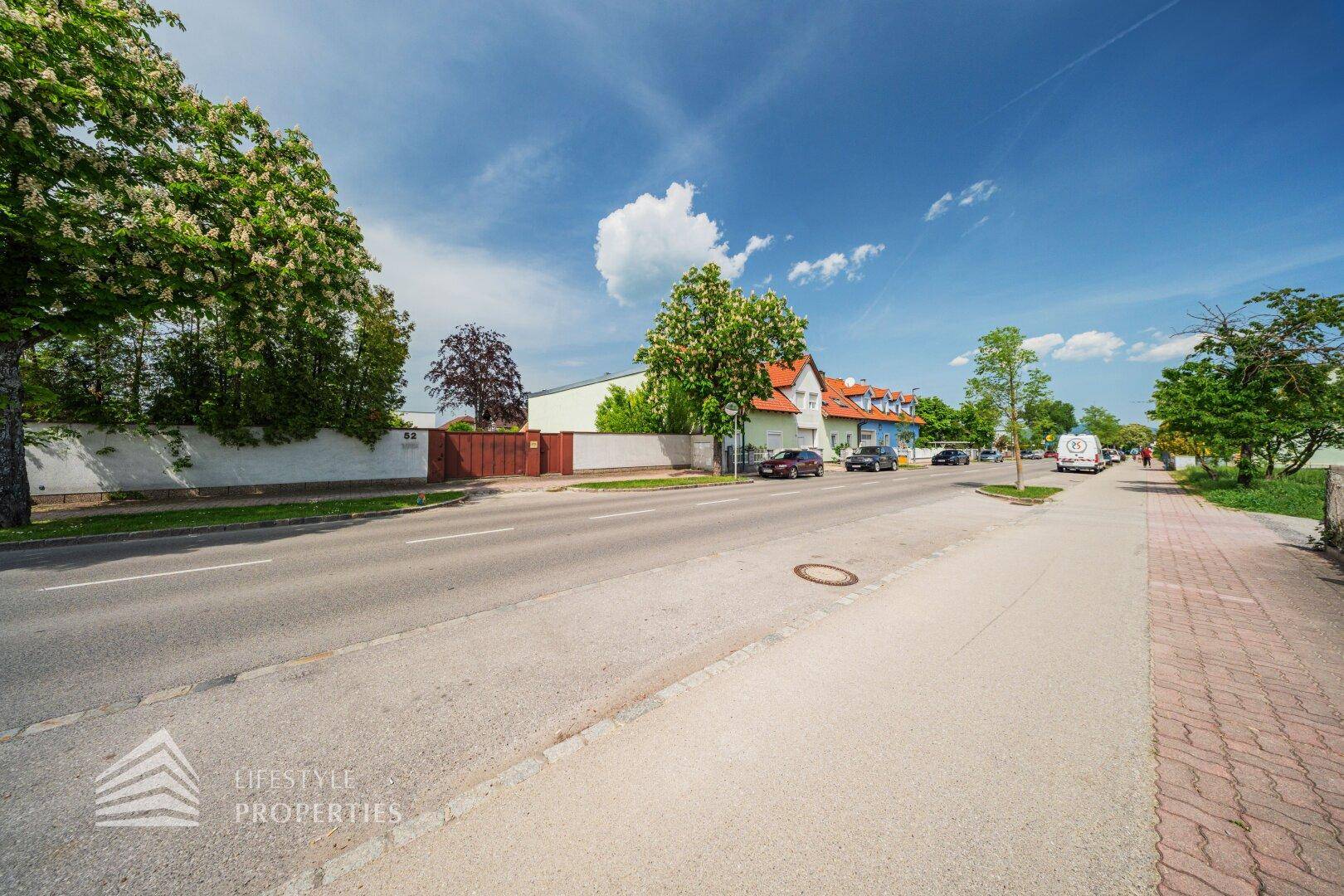 Weitläufige Straßenansicht mit Fahrbahn, Bürgersteig und Wohnhäusern in der Umgebung.