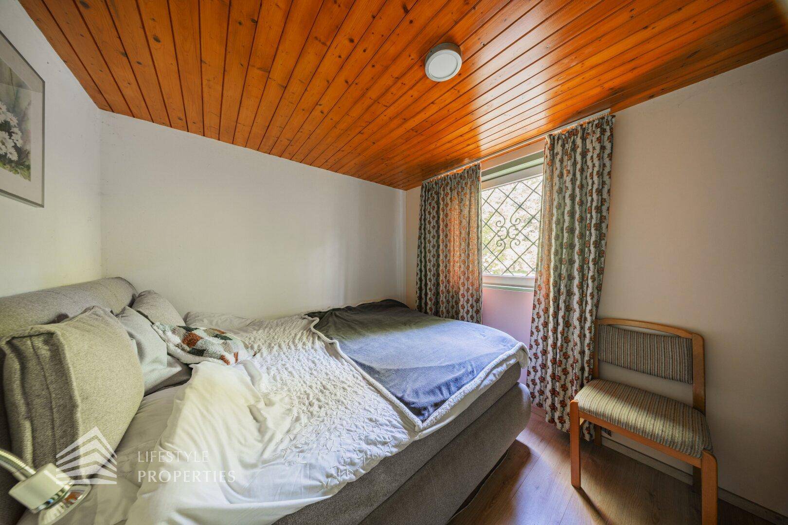 Schlafzimmer mit Doppelbett und Holzdecke, hell durch ein Fenster mit Gitter.