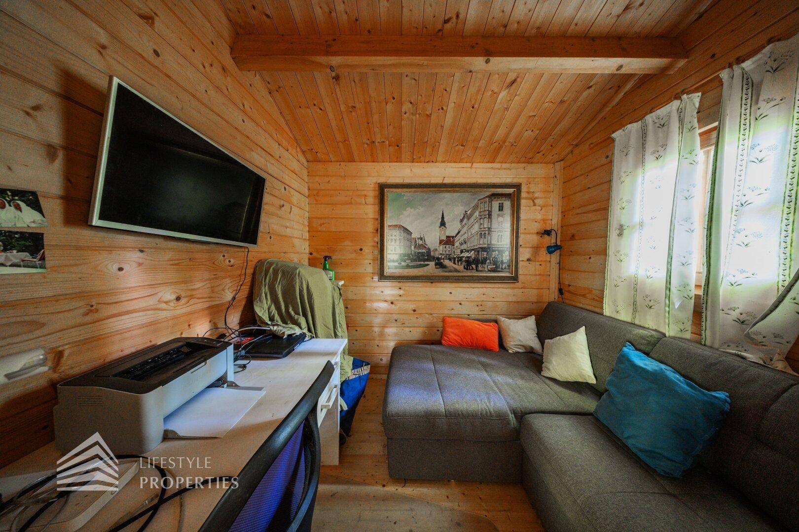 Wohnbereich in einer Holzhütte mit Sofa, Fernseher und Arbeitsplatz, umgeben von Holzvertäfelung.