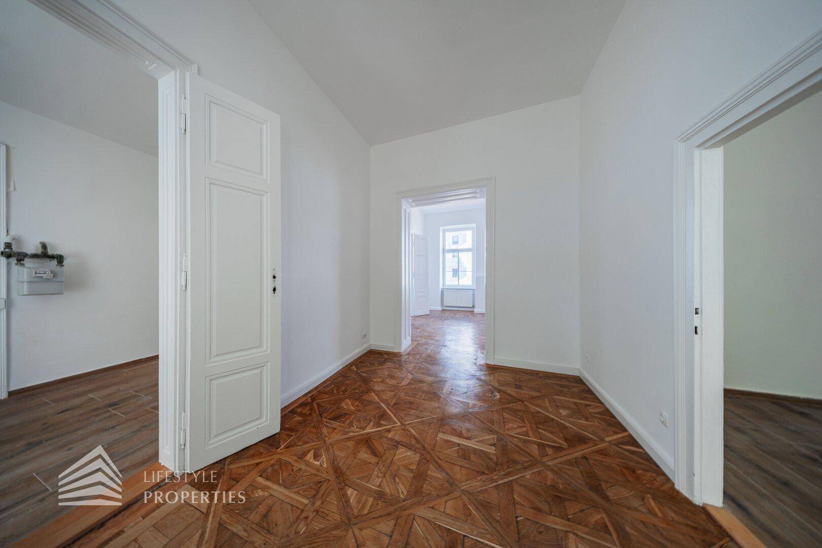Leerer Raum mit kunstvollem Parkettboden und weißen Wänden, der durch eine offene Tür zugänglich ist.
