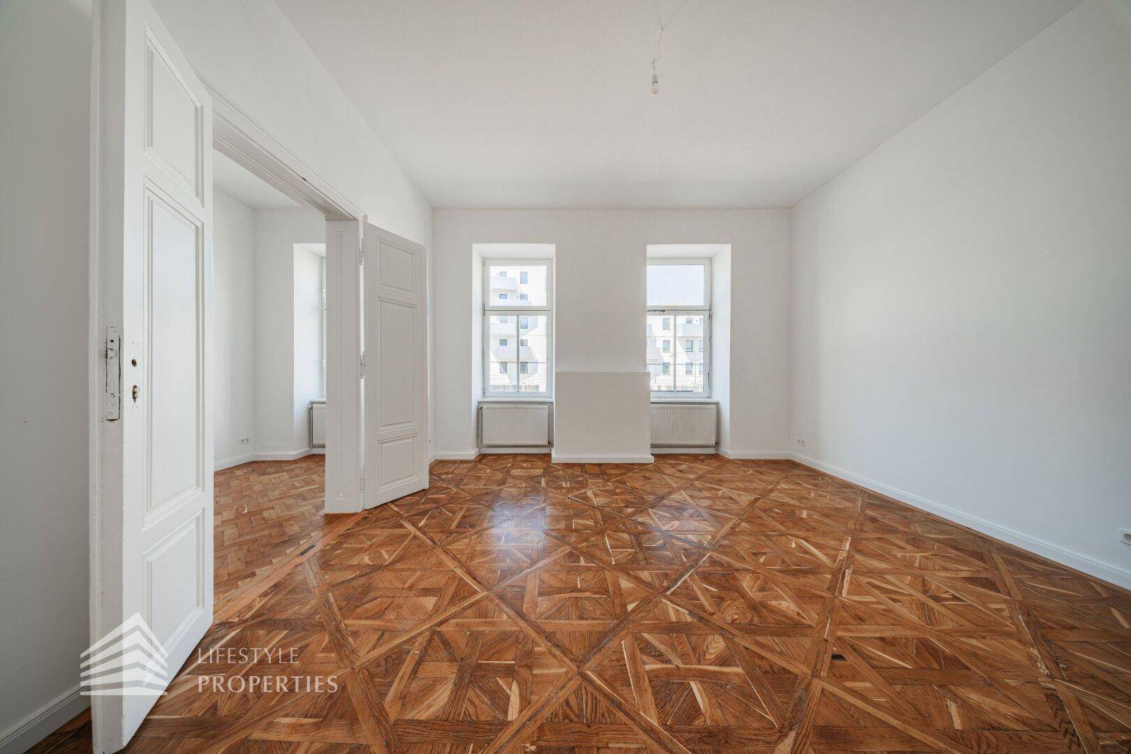 Helles, leeres Zimmer mit kunstvollem Parkettboden und zwei großen Fenstern.