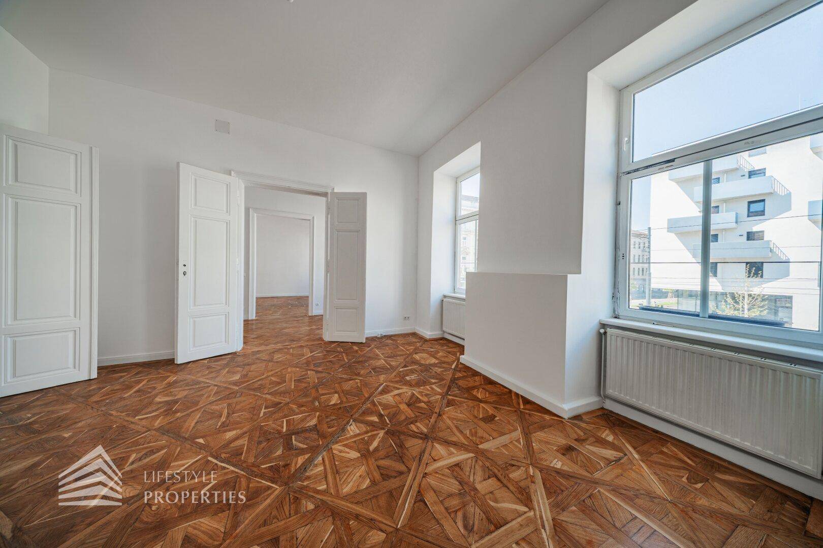 Geräumiges, leeres Zimmer mit kunstvollem Parkettboden und großen Fenstern, die viel Licht hereinlassen.