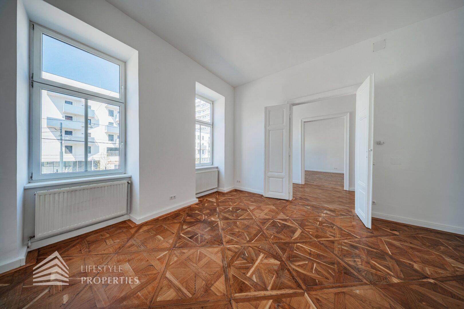 Helles, leeres Zimmer mit klassischem Parkett und hohen Fenstern, die einen Blick nach draußen bieten.