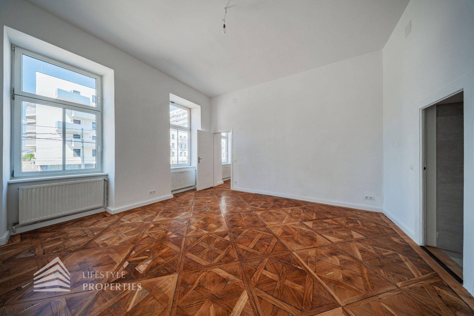 Helles, leeres Zimmer mit kunstvollem Parkettboden und großen Fenstern, die viel Licht hereinlassen.