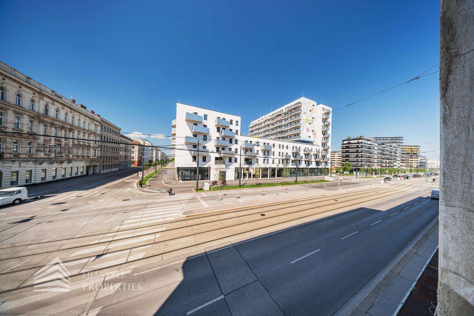 Weitläufige Straßenansicht mit modernen Gebäuden und Straßenbahnschienen unter blauem Himmel.