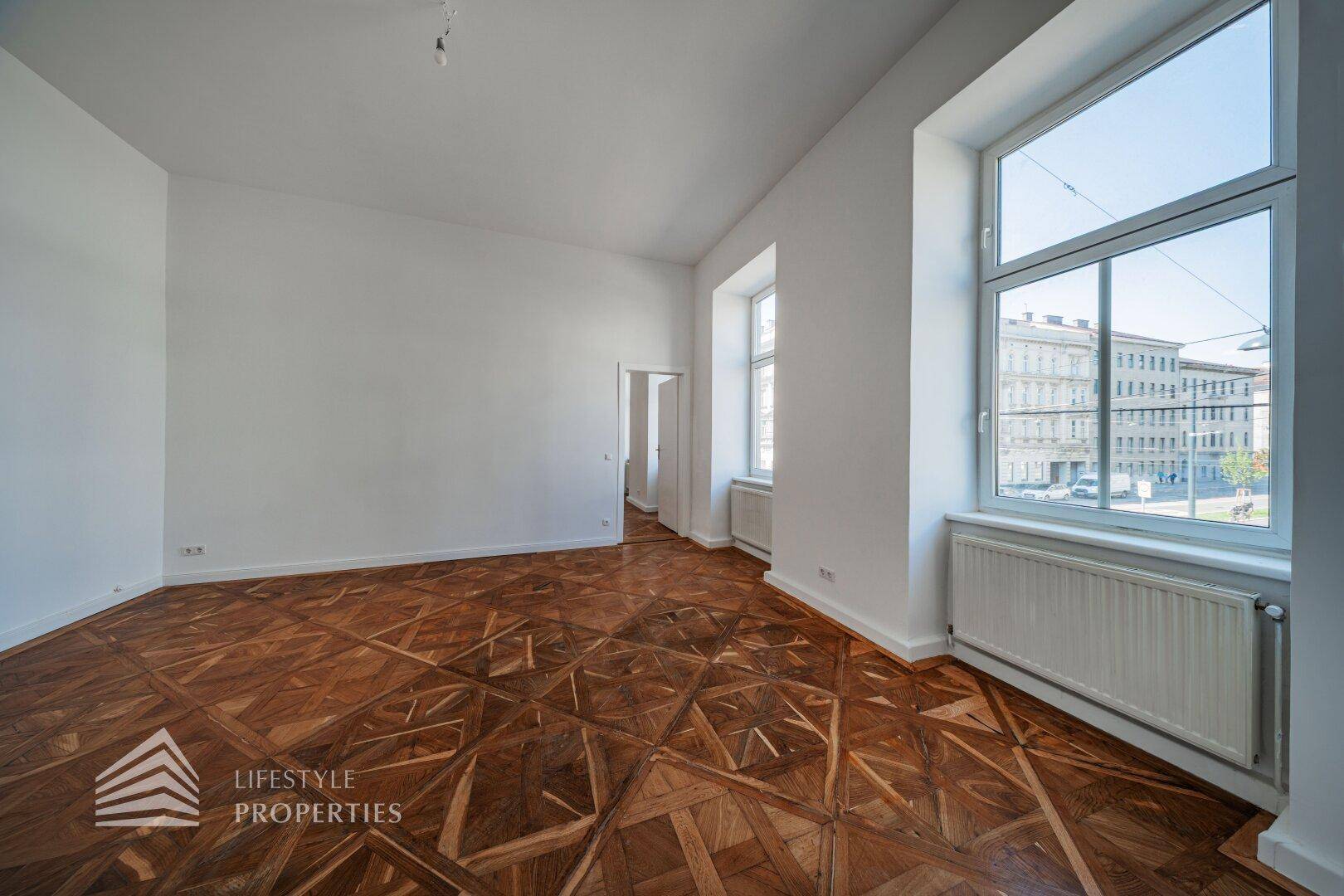 Geräumiges, leeres Zimmer mit kunstvollem Parkettboden und großen Fenstern mit Stadtblick.
