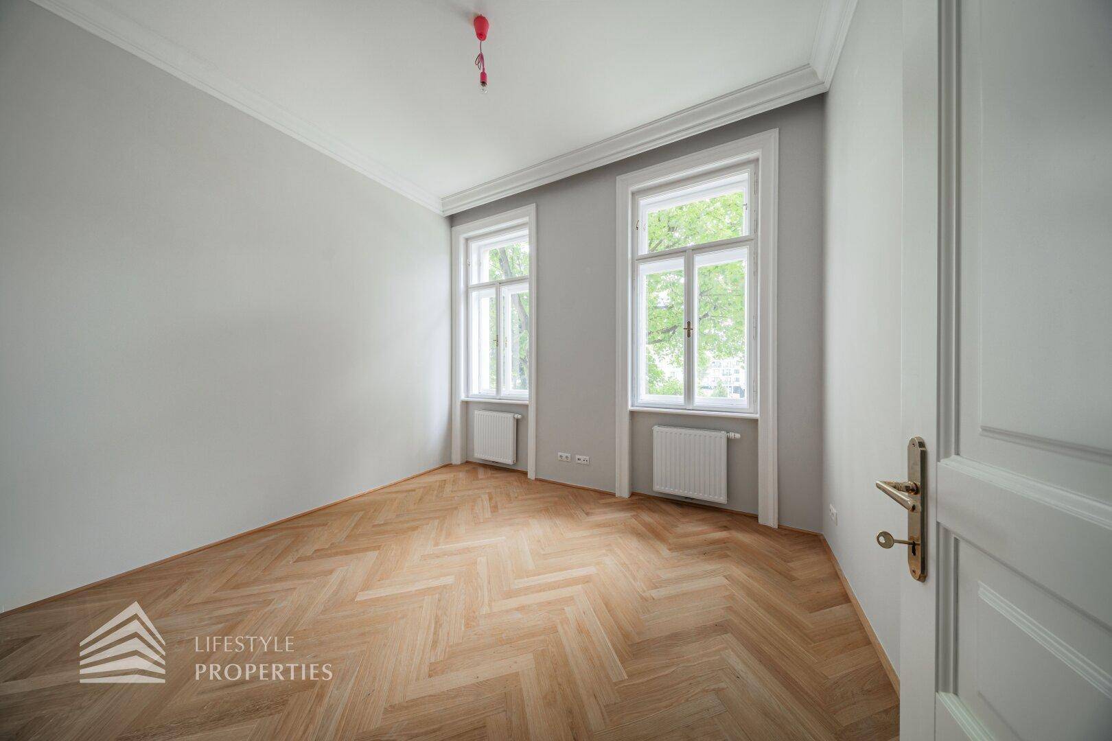 Helles Schlafzimmer mit Fischgrätparkett, hohen Fenstern und klassischer Deckenleiste.