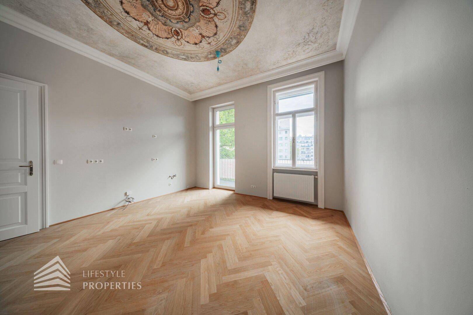 Geräumiger Raum mit Fischgrätparkett, hohen Decken und einem kunstvollen Stuckmedaillon an der Decke.