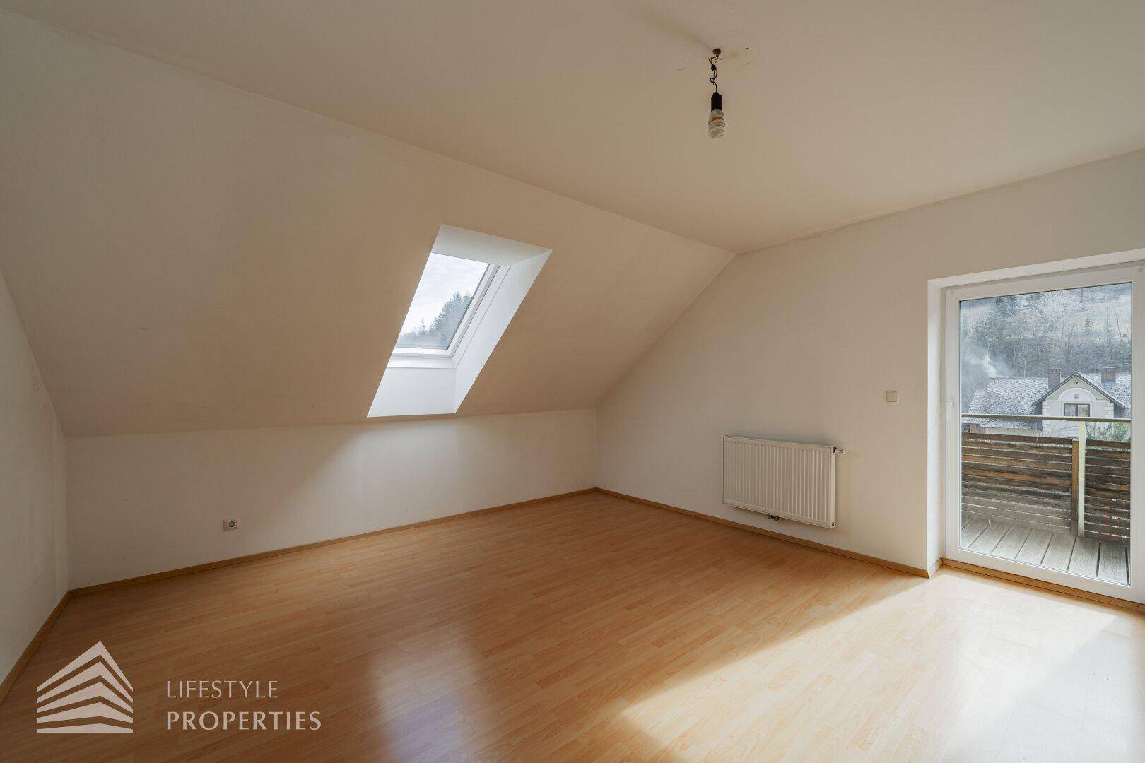 Heller Raum im Dachgeschoss mit Dachfenster, Holzboden und Zugang zum Balkon.