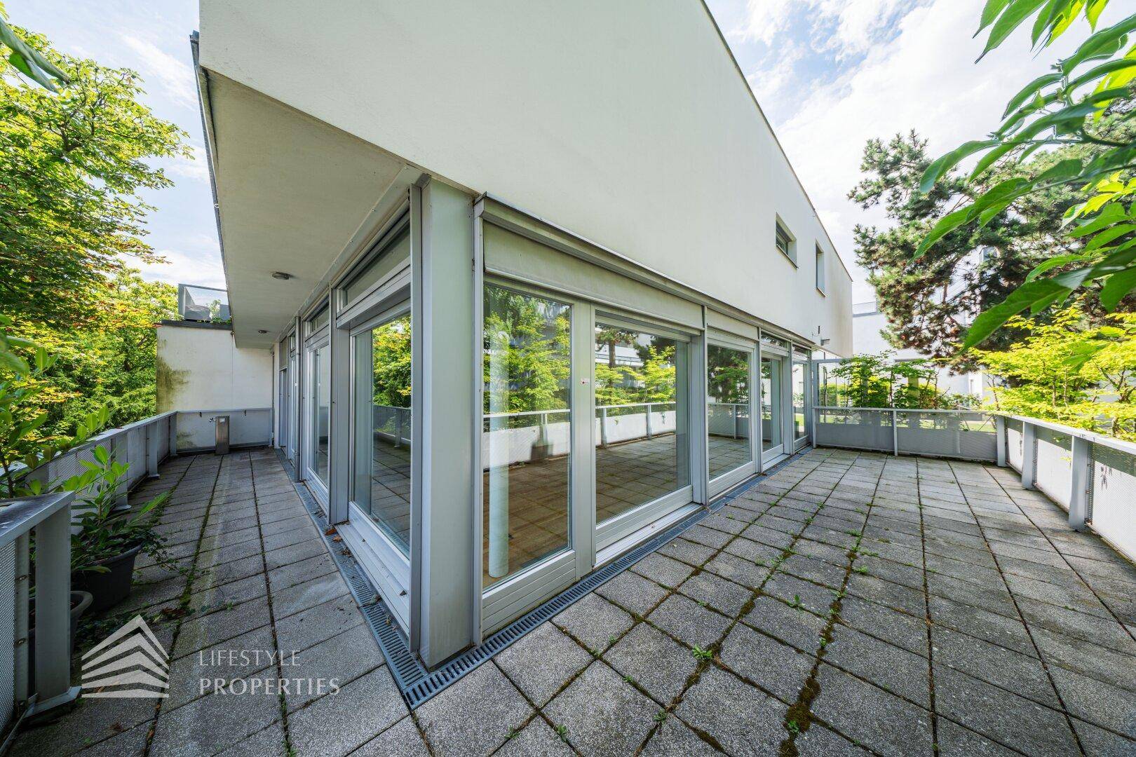 Eckterrasse mit Steinplatten und Blick ins Grüne, umgeben von Bäumen und Pflanzen.