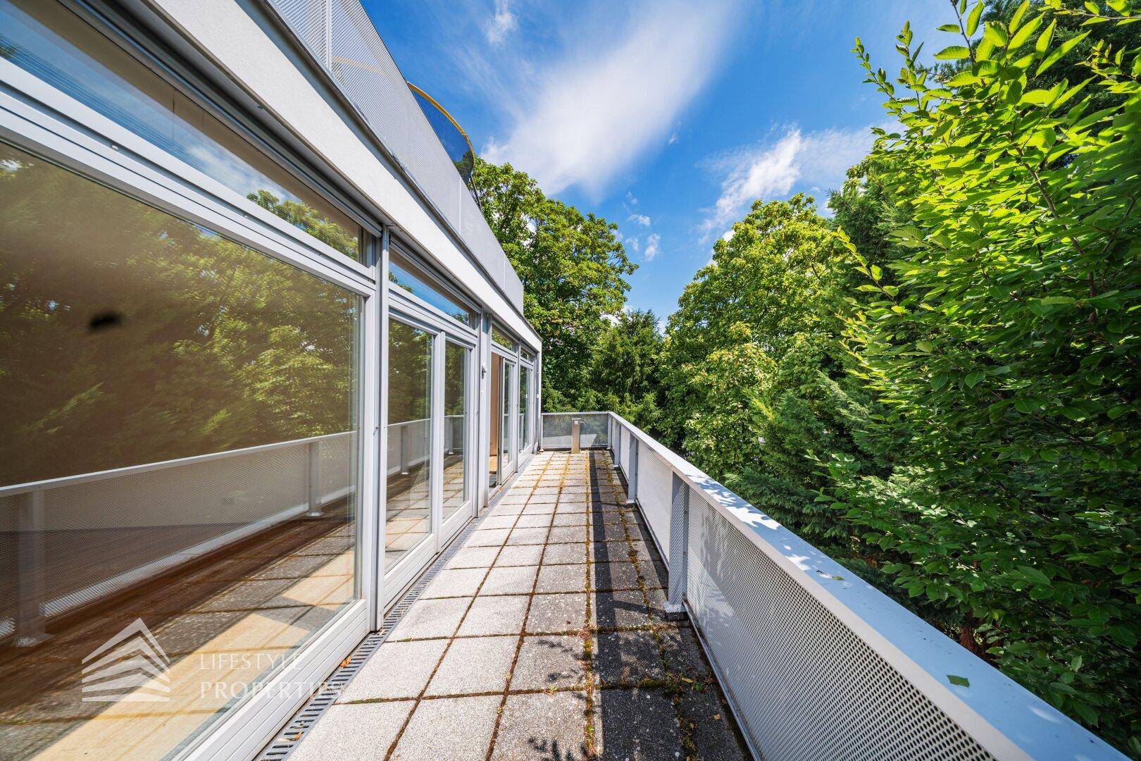 Sonnige Terrasse mit Steinplatten und moderner Brüstung, umgeben von üppigem Grün und blauem Himmel.
