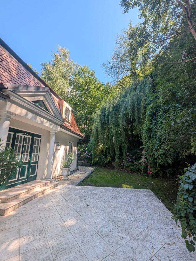 Große gepflasterte Terrasse neben dem Haus mit üppiger grüner Vegetation im Hintergrund.