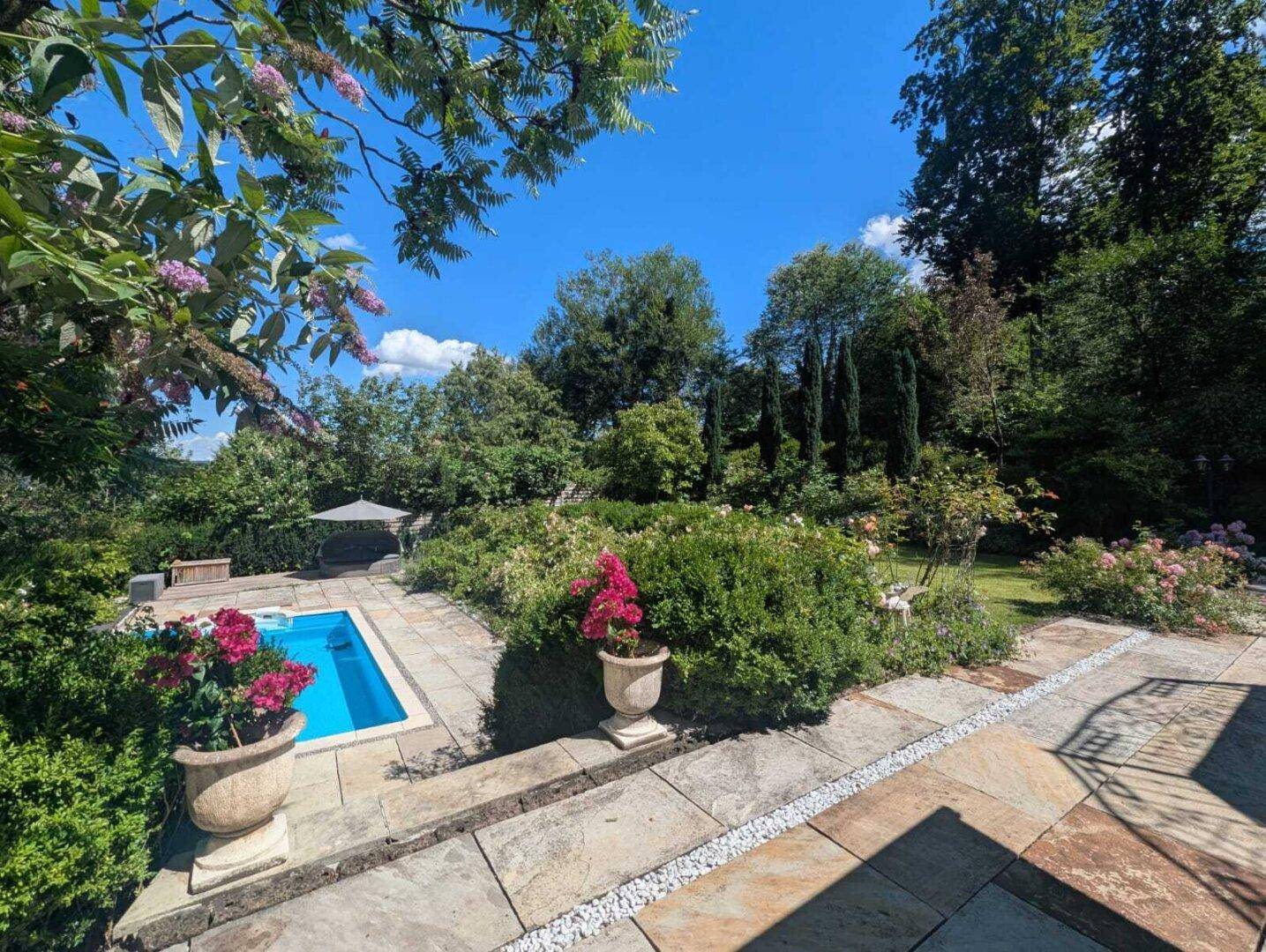 Gepflegter Garten mit Pool, üppiger Bepflanzung und hohen Bäumen unter blauem Himmel.