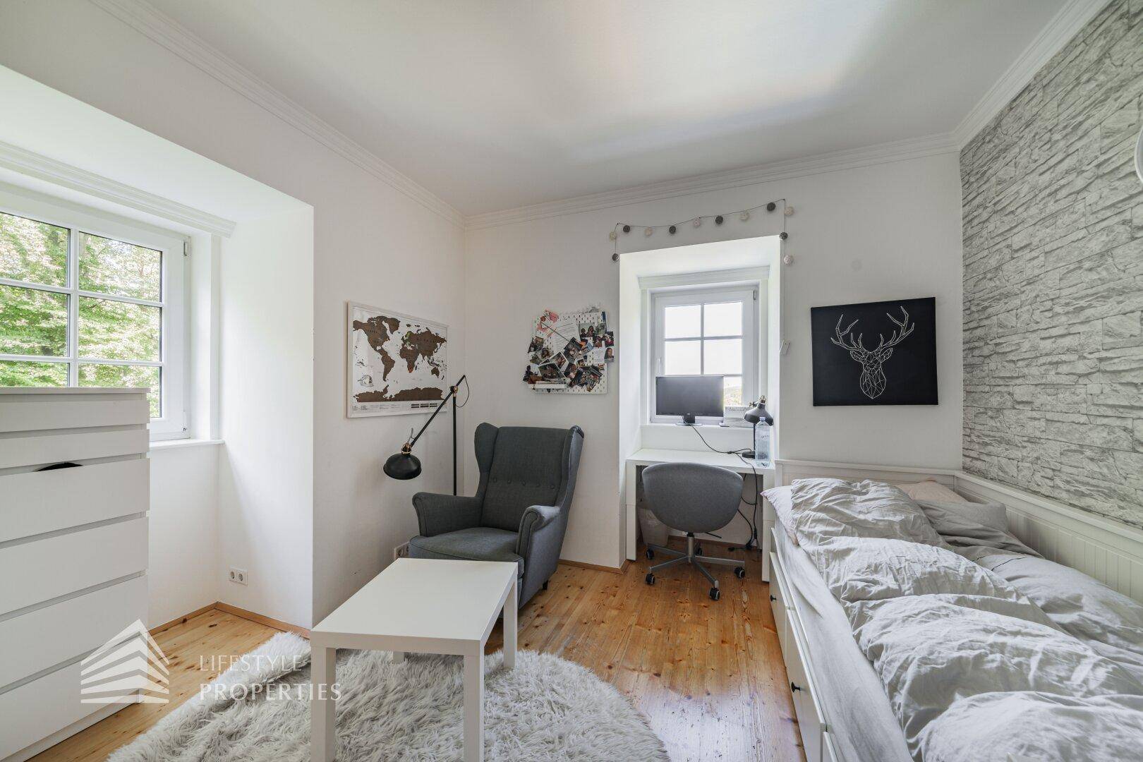 Freundliches Schlafzimmer mit Schreibtischbereich, Tagesbett und zwei Fenstern für viel Licht.