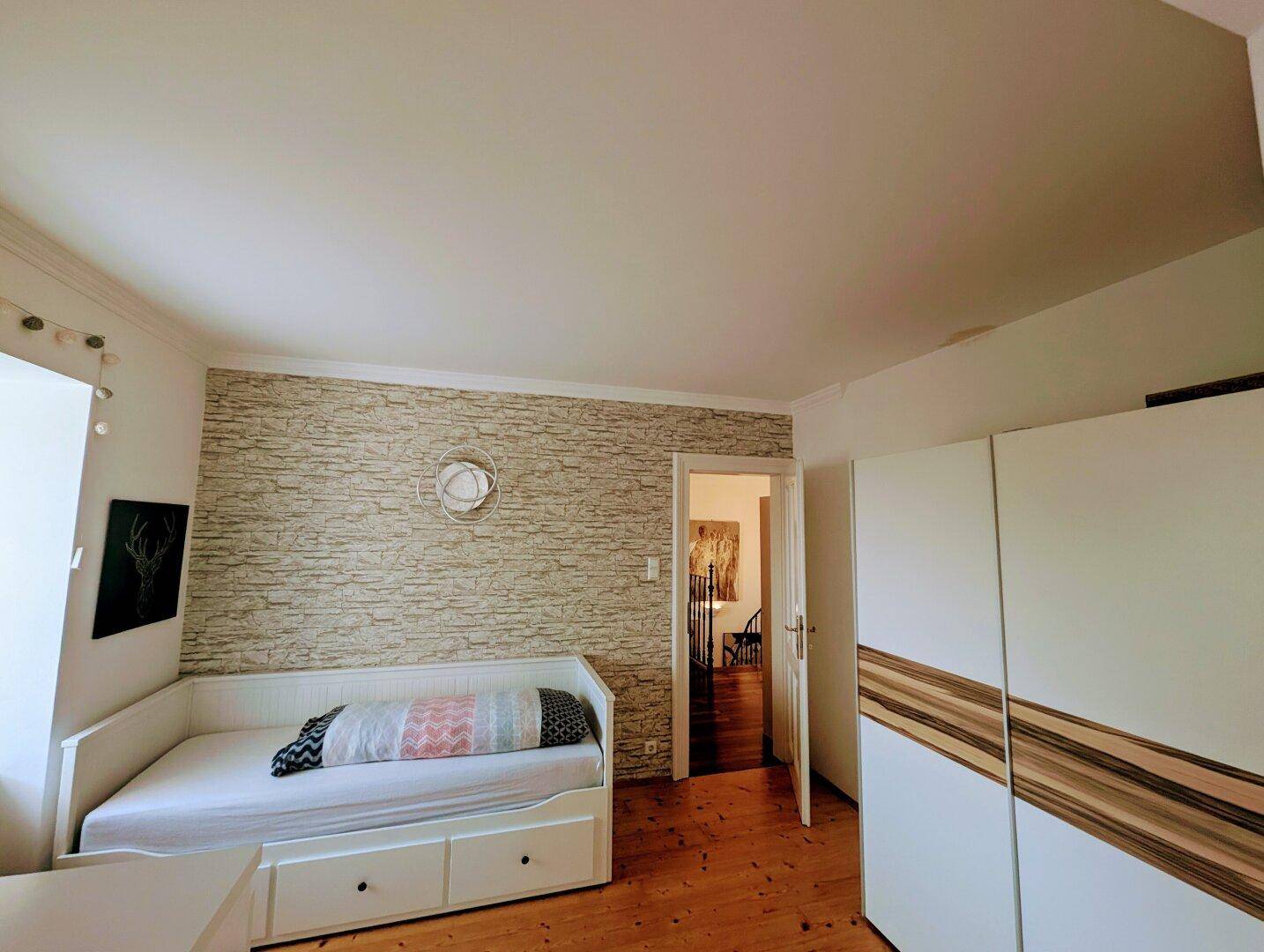 Helles Schlafzimmer mit Tagesbett, Kleiderschrank und dekorativer Steinoptik-Tapete.