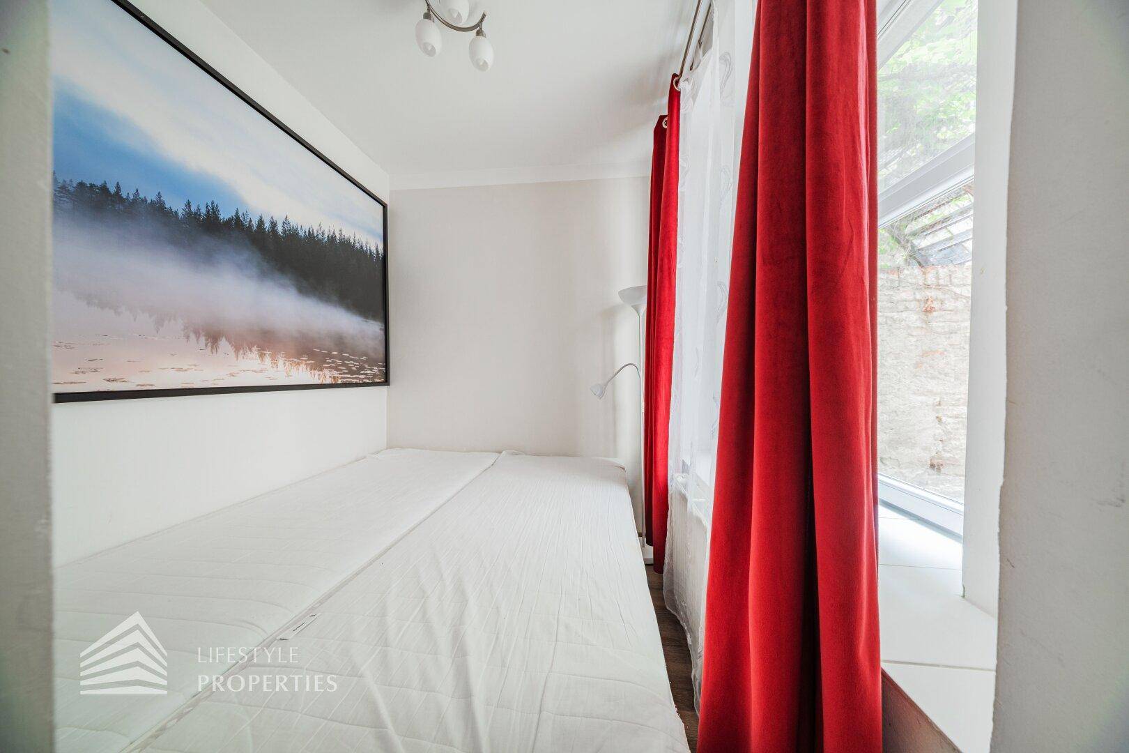Helles Schlafzimmer mit Einzelbett, großem Fenster und roten Vorhängen.