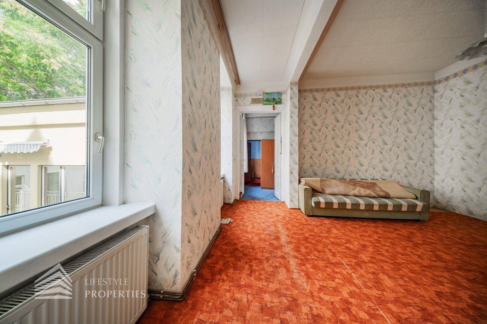 Helles Zimmer mit Heizkörper unter dem Fenster und Blick auf die Umgebung, in ursprünglichem Zustand.