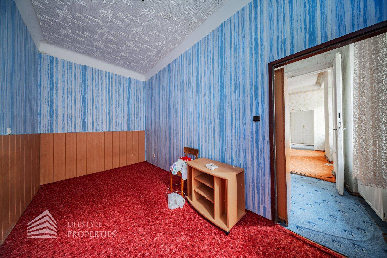 Flur mit blauer Tapete und rotem Teppich, der zu weiteren Räumen führt, mit einfacher Einrichtung.