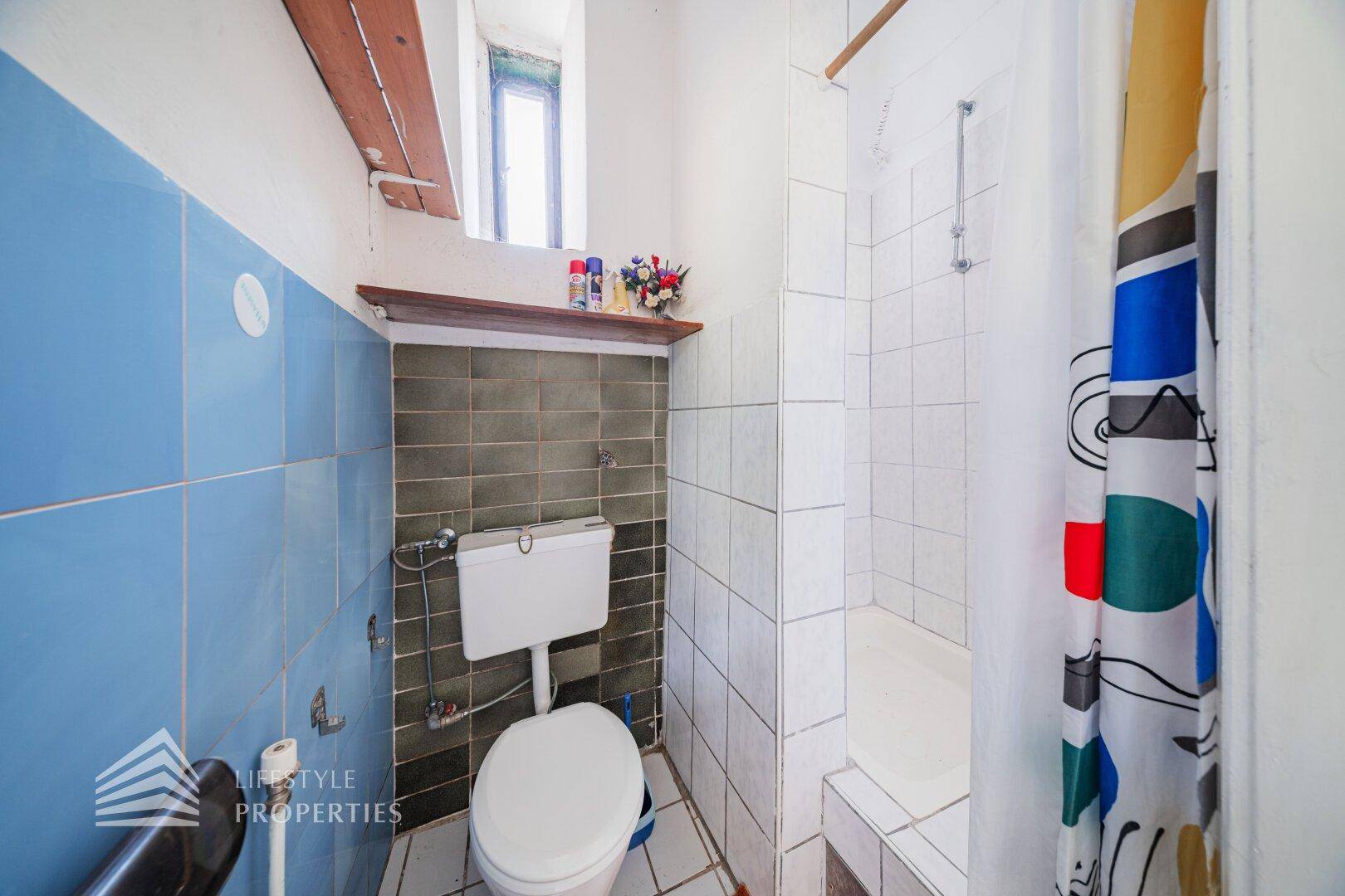 Kleines Badezimmer mit WC, Dusche und blauen sowie weißen Fliesen, im Stil der 80er Jahre.