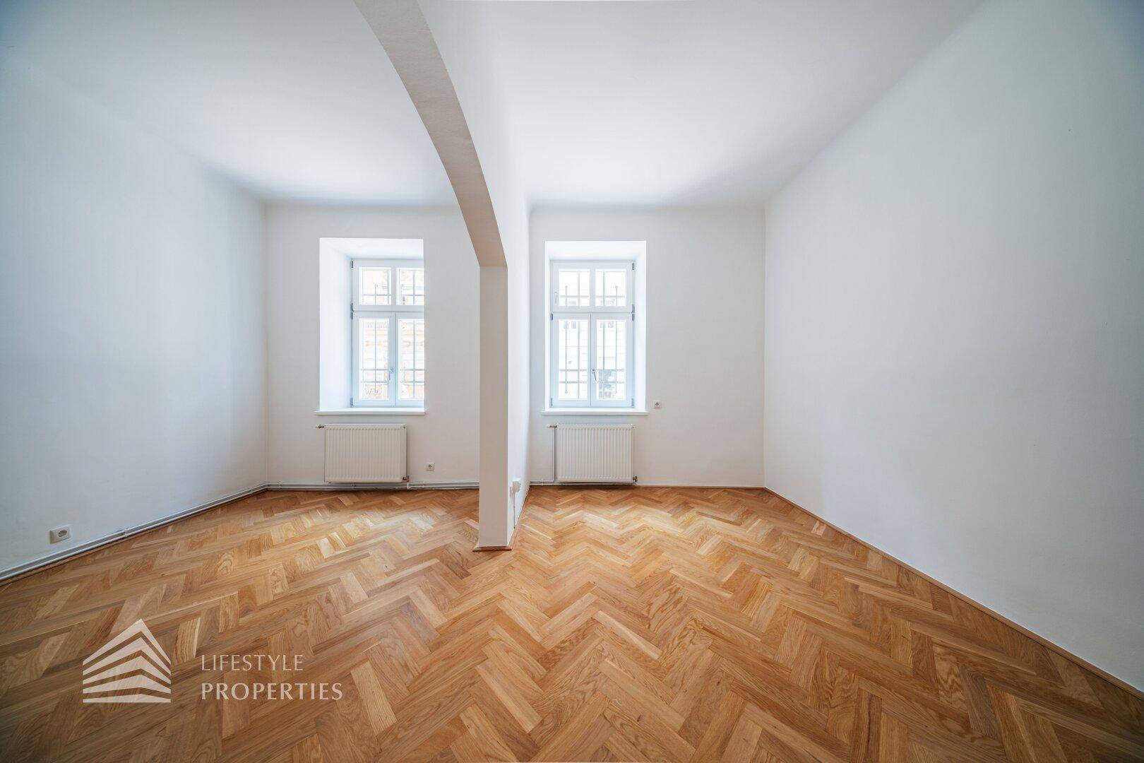 Geräumiges, helles Zimmer mit klassischem Fischgrätparkett und zwei großen Fenstern.