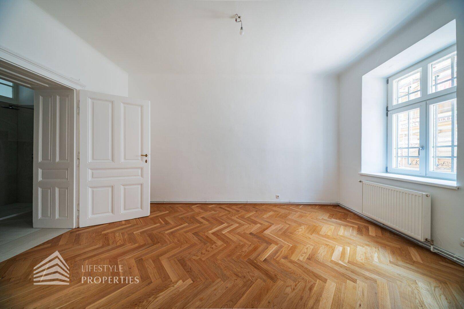 Lichtdurchflutetes Zimmer mit Fischgrätparkett und einem großen Fenster, das viel Tageslicht hereinlässt.