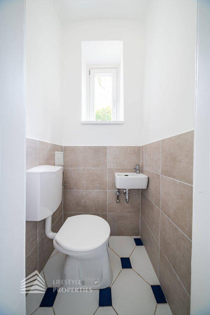 Separates WC mit Fenster, Wandtoilette und kleinem Waschbecken, ausgestattet mit Fliesen.