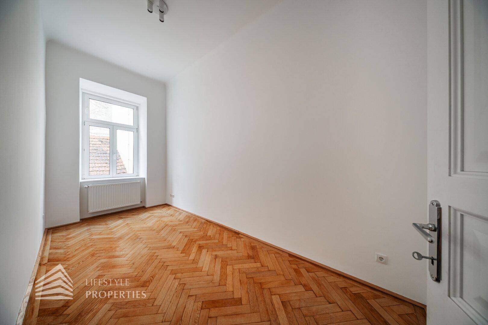 Unmöbliertes Zimmer mit klassischem Fischgrätparkett und einem Fenster, das den Raum erhellt.