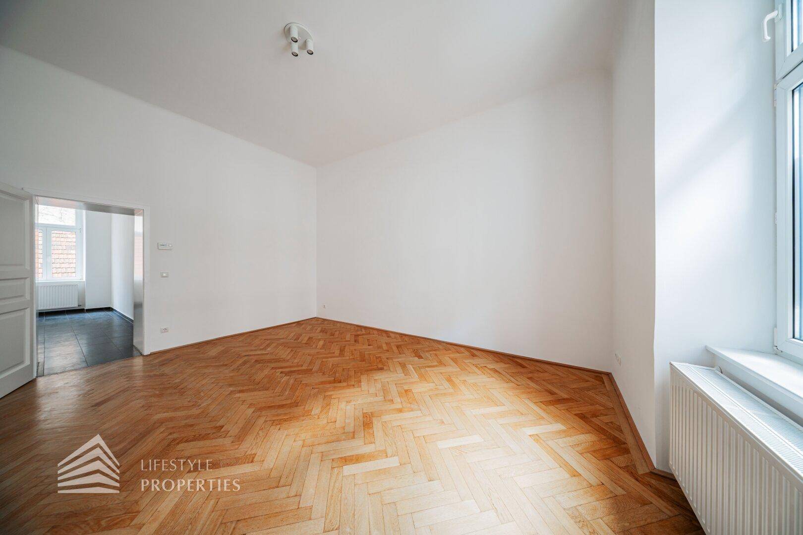 Geräumiges, unmöbliertes Zimmer mit Fischgrätparkett und Zugang zu einem weiteren Raum.