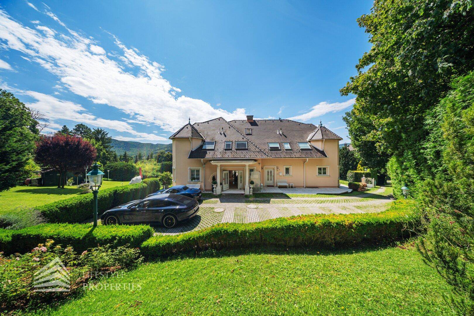 Die imposante Villa mit weitläufiger Auffahrt, umgeben von einem gepflegten Garten und alten Bäumen.