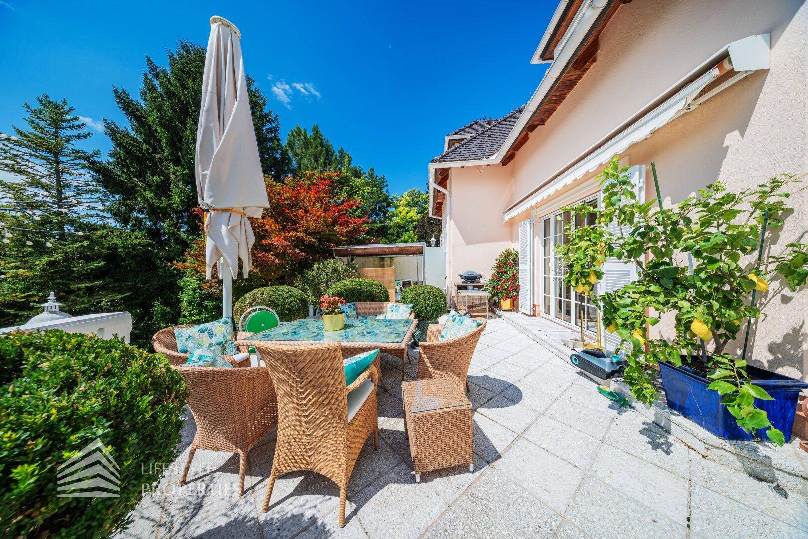 Eine sonnige Terrasse mit Rattanmöbeln, Sonnenschirm und Pflanzen, ideal für entspannte Stunden im Freien.