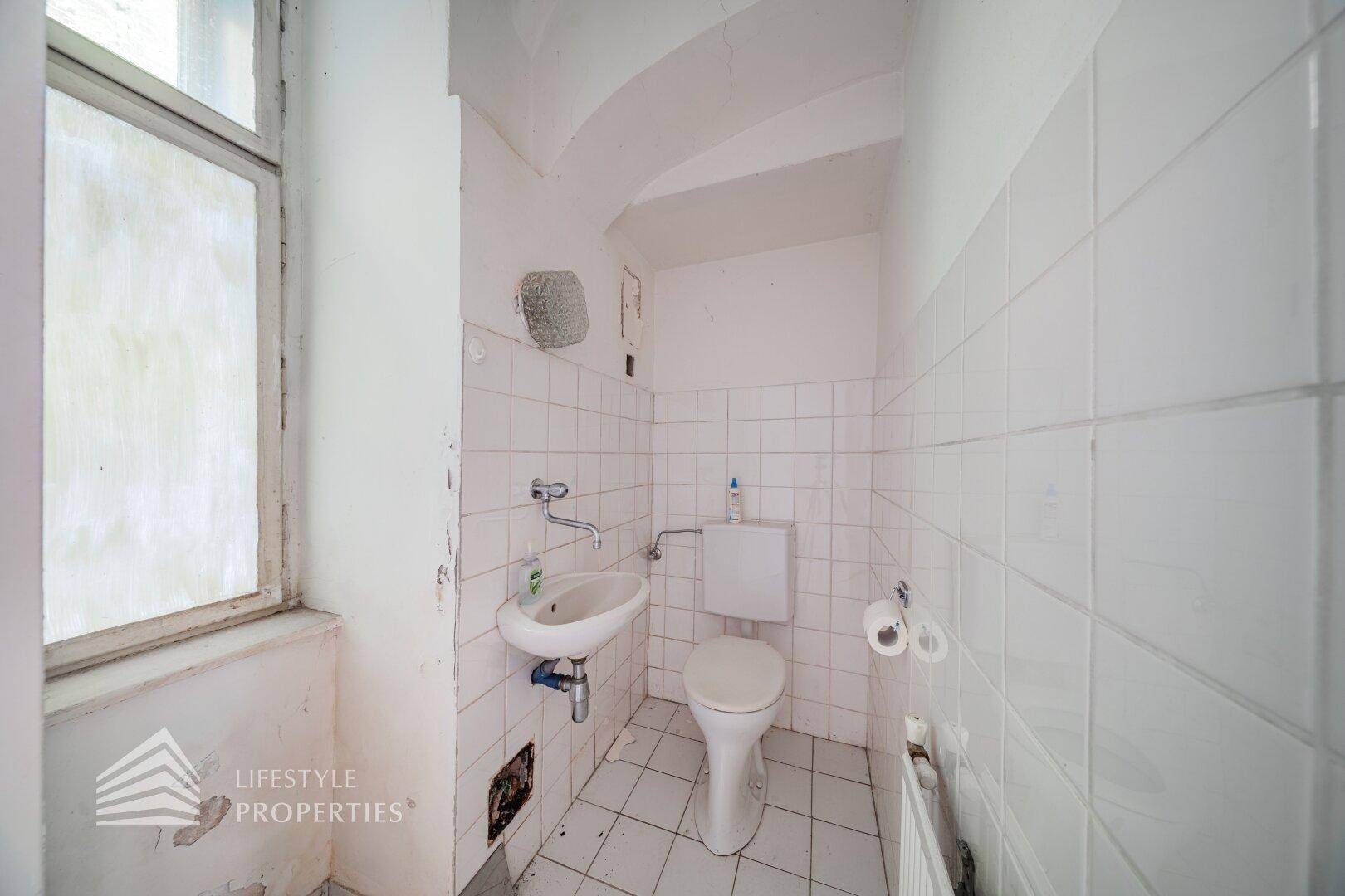 Einfache Gästetoilette mit Fenster, Waschbecken und WC, gefliest in Weiß.