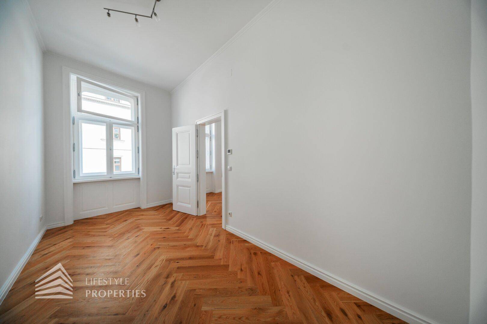 Helles Zimmer mit Fischgrätparkett und großen Fenstern, das viel Tageslicht hereinlässt.