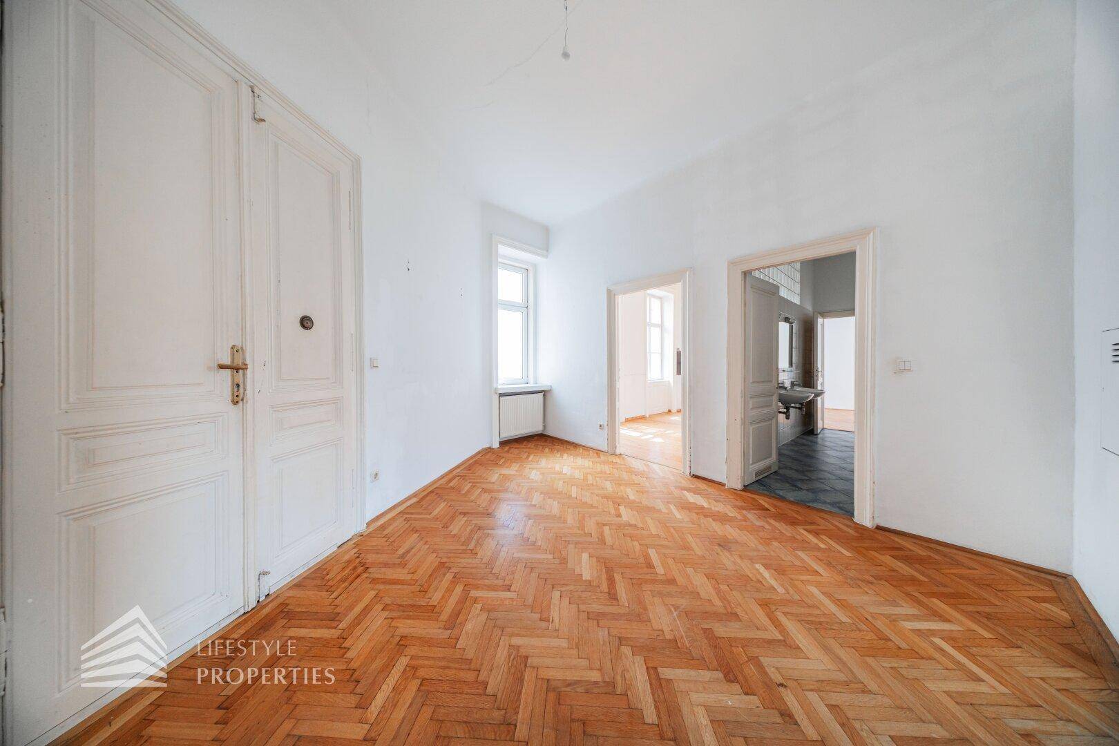 Leeres Zimmer mit Fischgrätparkett und hohen Flügeltüren, das auf eine Renovierung wartet.