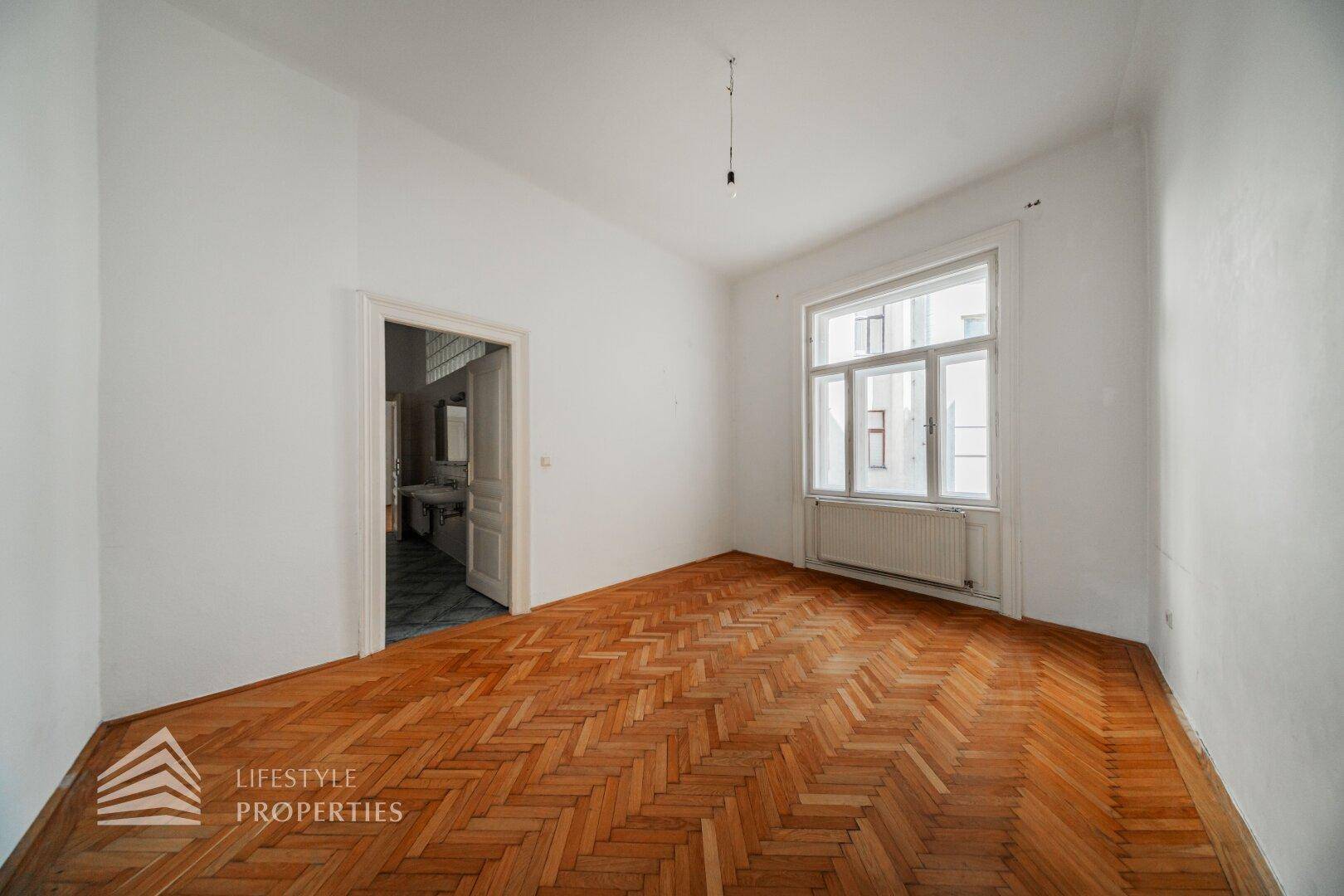 Leeres Zimmer mit Fischgrätparkett und einem Fenster, das in einen weiteren Raum führt.