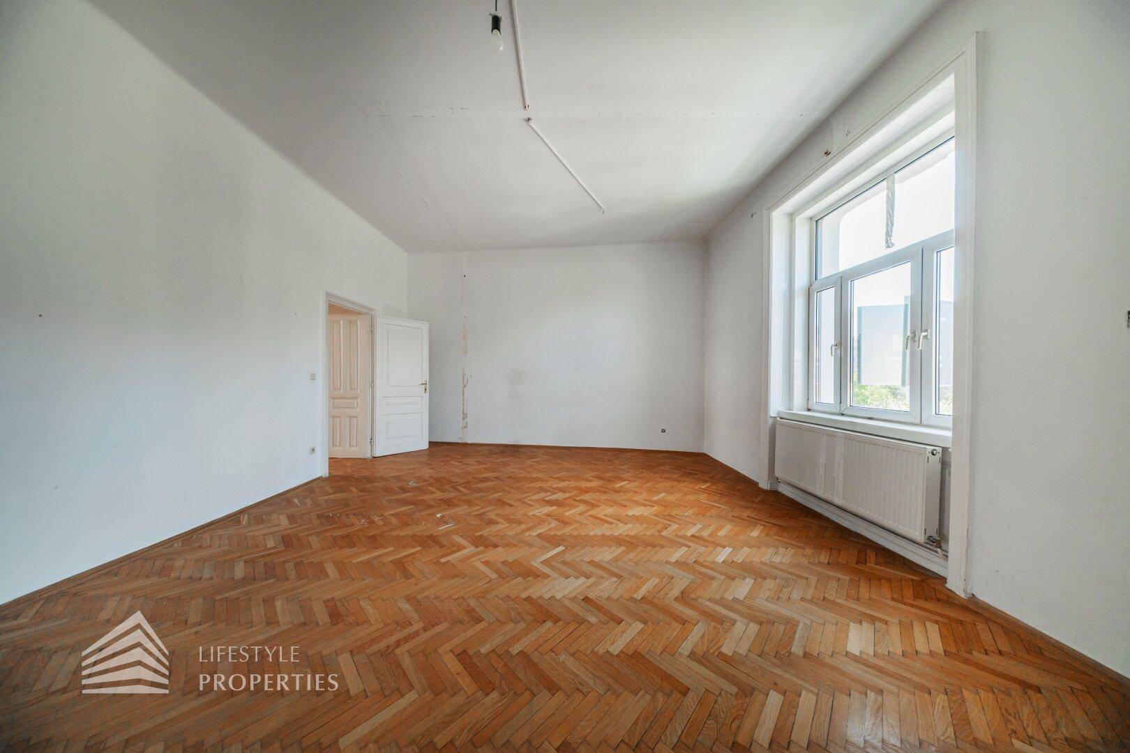 Helles Zimmer mit Fischgrätparkett und großen Fenstern, die viel Tageslicht hereinlassen.