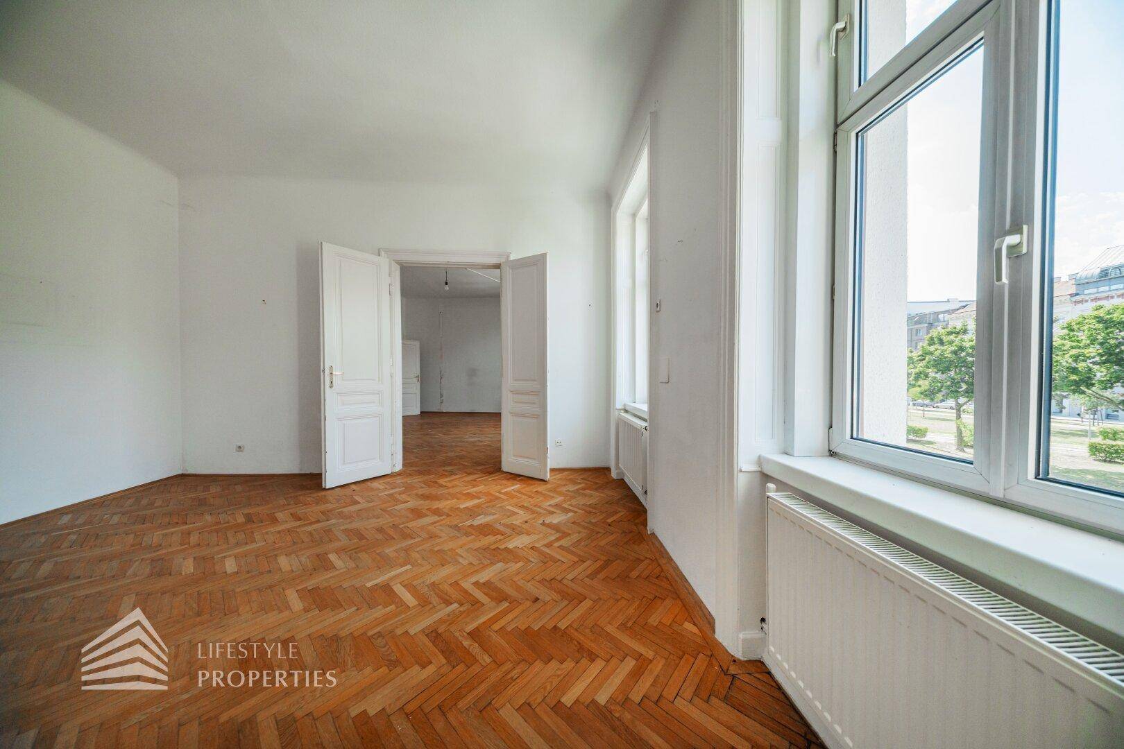 Geräumiges Zimmer mit klassischem Fischgrätparkett und Blick ins Grüne durch große Fenster.