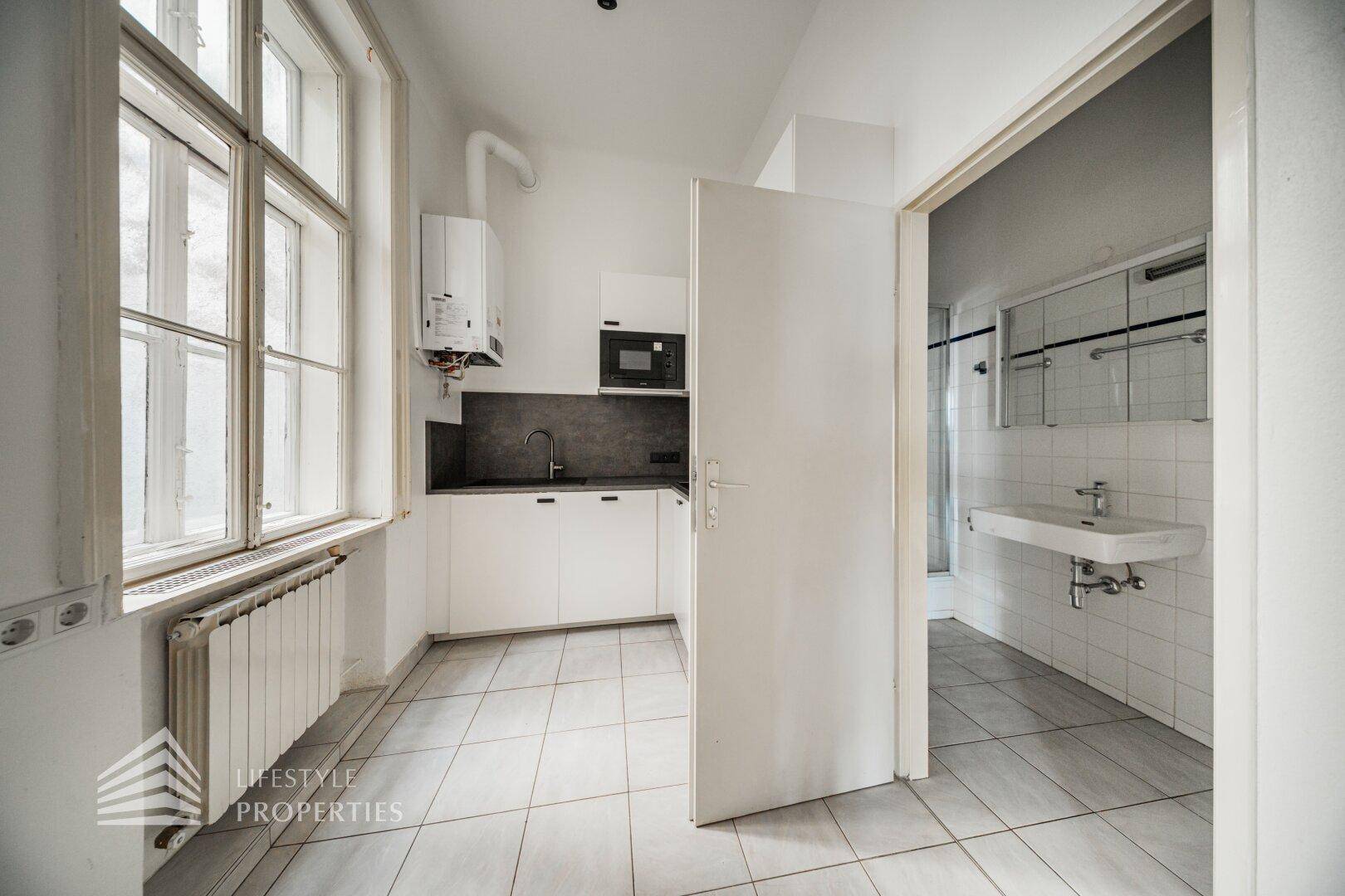 Kleine Küche mit weißen Schränken, Fliesenboden und Blick in das angrenzende Badezimmer.