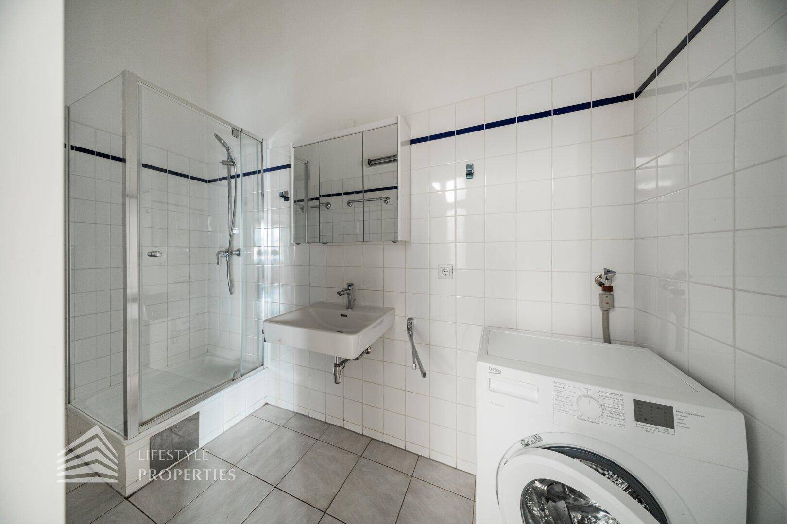 Badezimmer mit Duschkabine, Waschmaschine und weißen Fliesen mit blauen Akzenten.