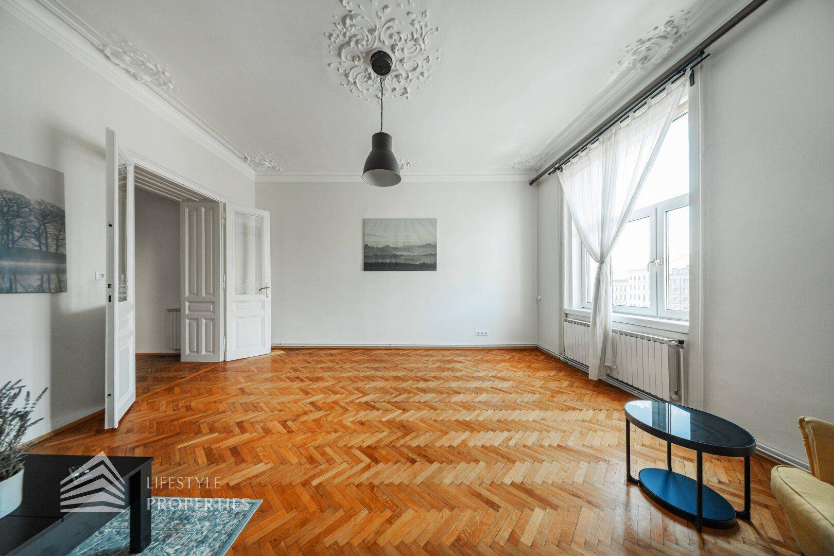 Leeres Wohnzimmer mit elegantem Fischgrätparkett und hohen Decken mit Stuckverzierungen.