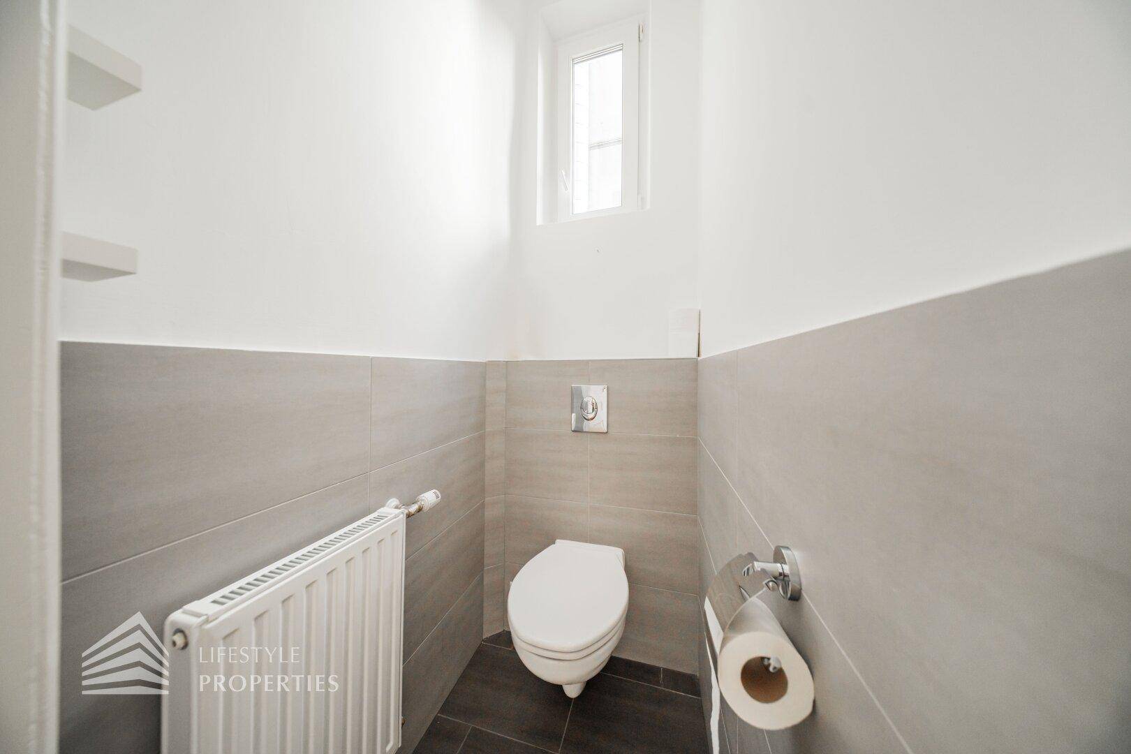 Separates WC mit wandhängender Toilette, kleinem Fenster und modernen Fliesen.